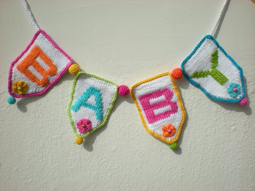 Gestrickte Girlande mit vier weißen Wimpeln, die die Buchstaben BABY in bunten Farben zeigen, verziert mit kleinen Blumen und Kugeln.