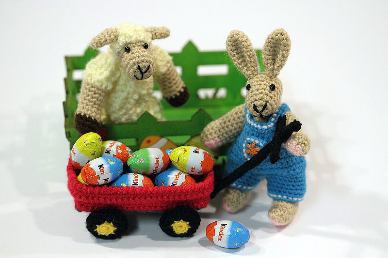 Roter Häkel-Bollerwagen mit bunten Eiern, gezogen von einem Amigurumi-Hasen; dahinter ein Amigurumi-Schaf vor grünem Zaun.