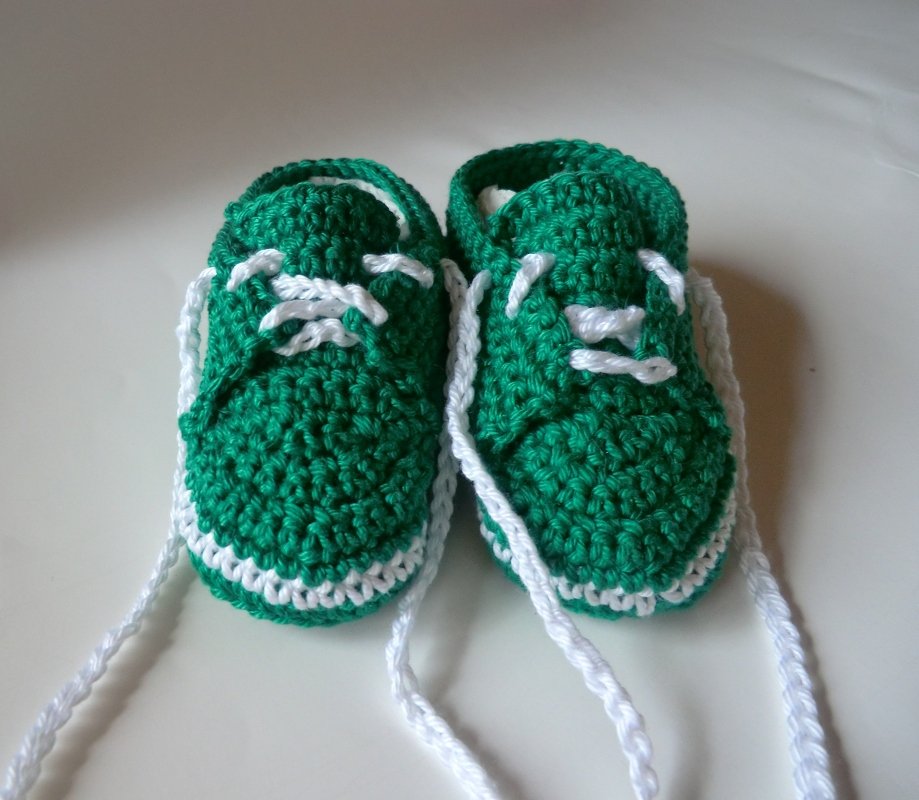 Zwei gehäkelte Babyschuhe in Grün mit weißen Schnürsenkeln und weißem Sohlenrand auf hellem Hintergrund.