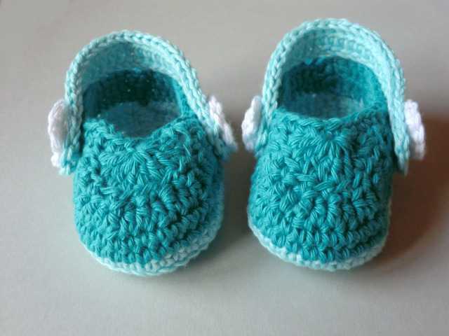 Paar türkisfarbener gehäkelter Babyschuhe mit hellblauen und weißen Details auf neutralem Hintergrund