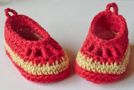 Zwei gehäkelte Babyschuhe in Rot und Gelb auf hellem Hintergrund, seitlich angeordnet