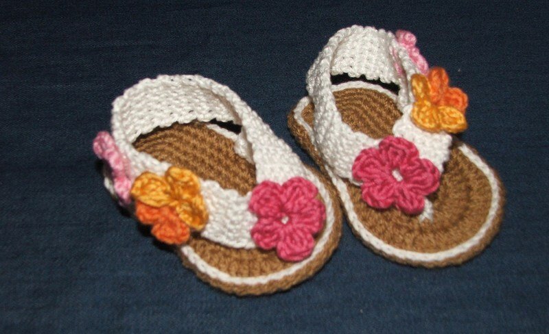 Gehäkelte Babysandalen mit brauner Sohle, weißen Riemen und rosa und orangefarbenen Blüten auf dunklem Untergrund.