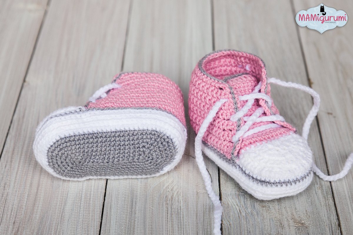 Gehäkelte Babyschuhe in Rosa, Weiß und Grau im Sneaker‑Stil auf hellem Holz; ein Schuh seitlich, der andere mit der Sohle nach oben.