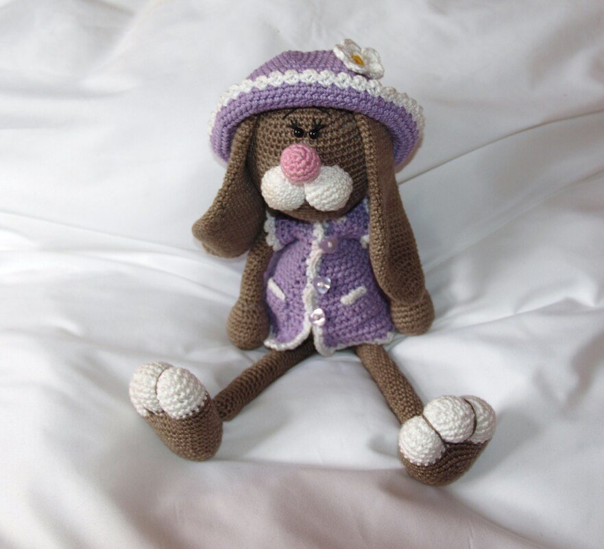Gehäkelter brauner Amigurumi-Hase mit lila Hut und Kleid auf weißer Oberfläche.