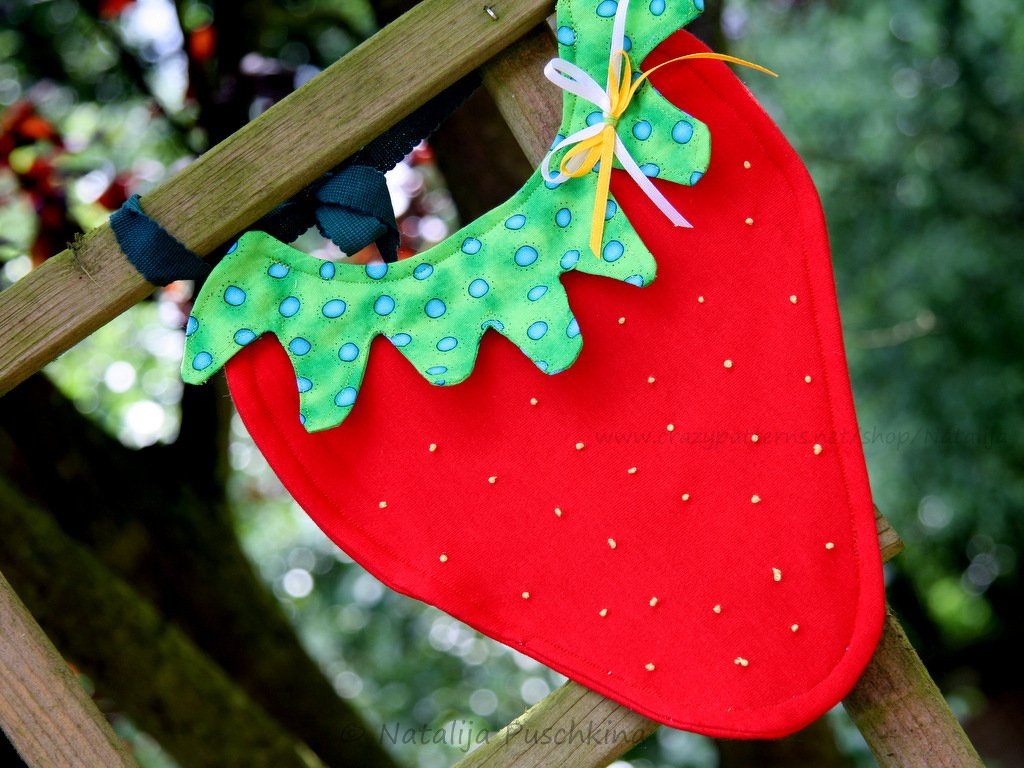 Rotes Babylätzchen in Erdbeerform mit grünem, gepunktetem Blatt, an einer Holzleiter hängend.