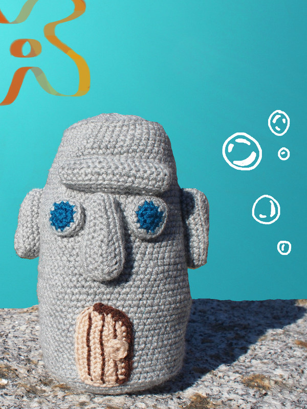 Gehäkelte graue Amigurumi-Figur mit blauen Augen und kleiner brauner Tür auf steinigem Untergrund vor türkisem Hintergrund.