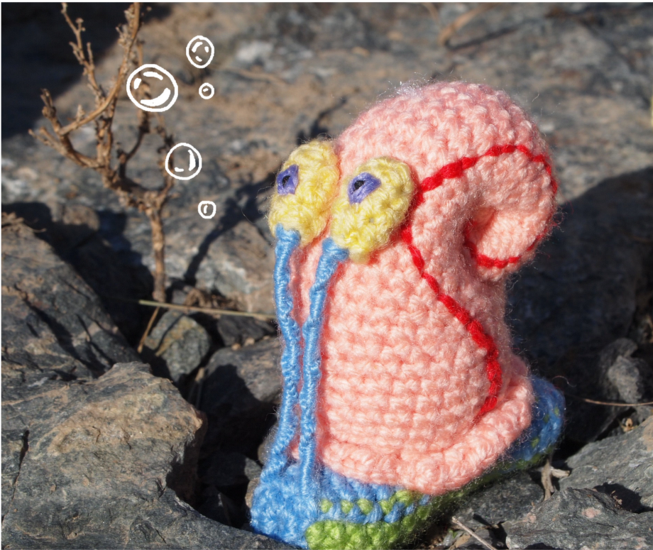 Gehäkelte Amigurumi‑Schnecke mit rosa Gehäuse, gelben Augen und blauen Fühlern auf Felsen