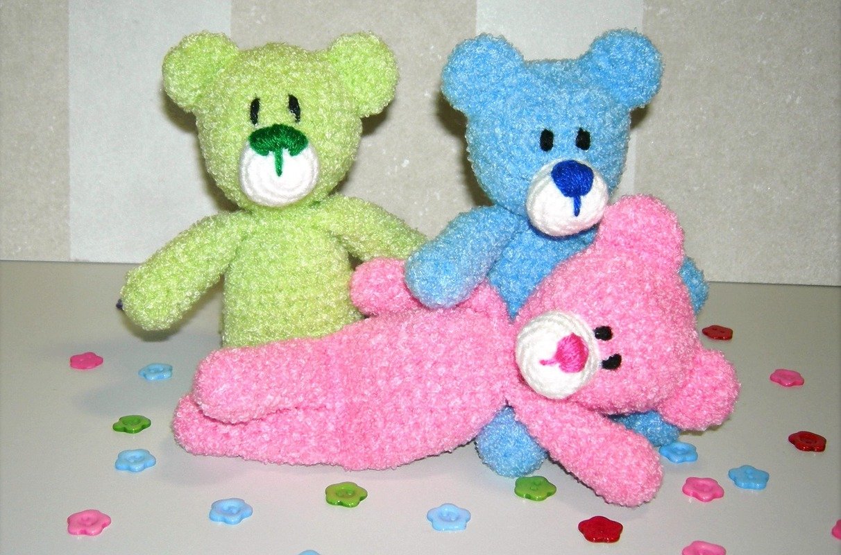 Drei gehäkelte Amigurumi-Teddys (grün, blau, rosa) auf heller Oberfläche, umgeben von bunten Blumenknöpfen.
