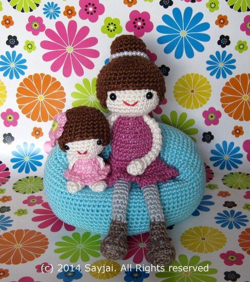 Two crocheted amigurumi dolls, a larger one in purple and a smaller one in pink, seated on a blue pouf with a floral background.
