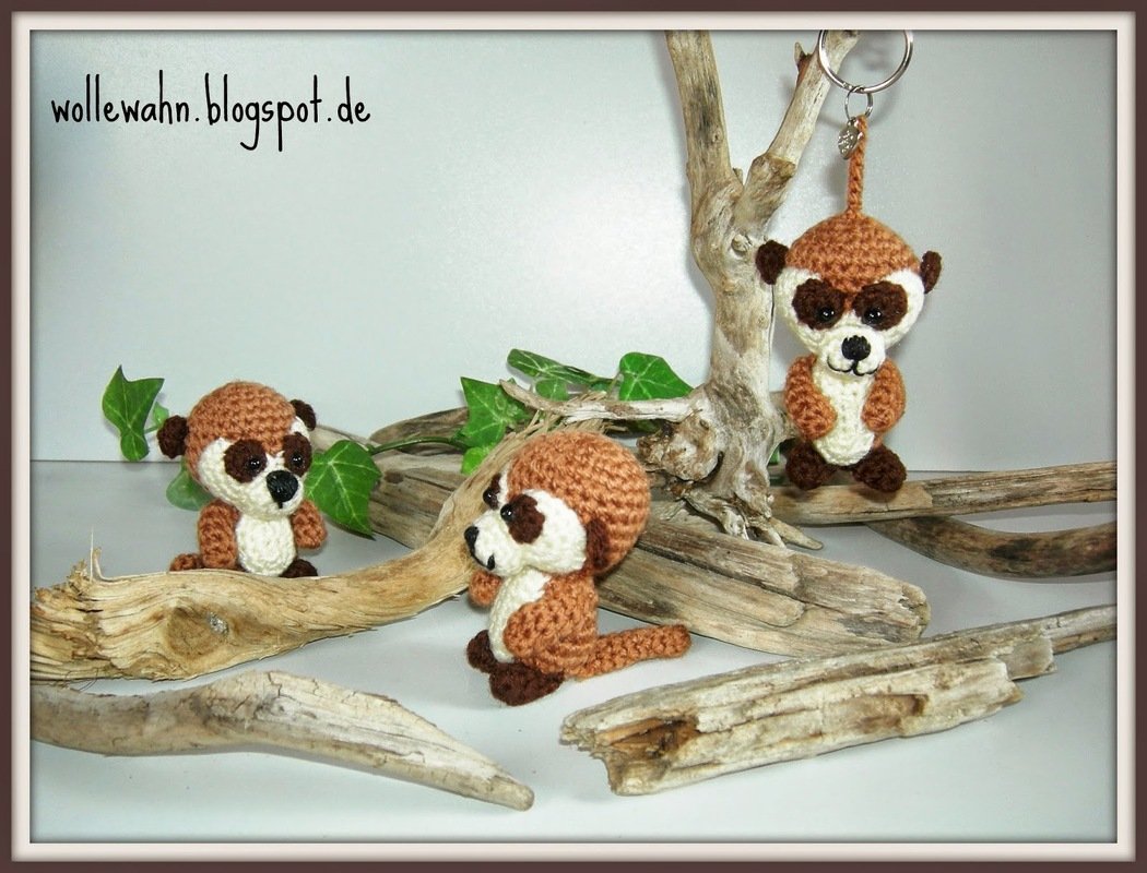 Drei gehäkelte Amigurumi-Erdmännchen in Braun und Beige, eines mit Schlüsselring, arrangiert auf Treibholz mit grünen Blättern