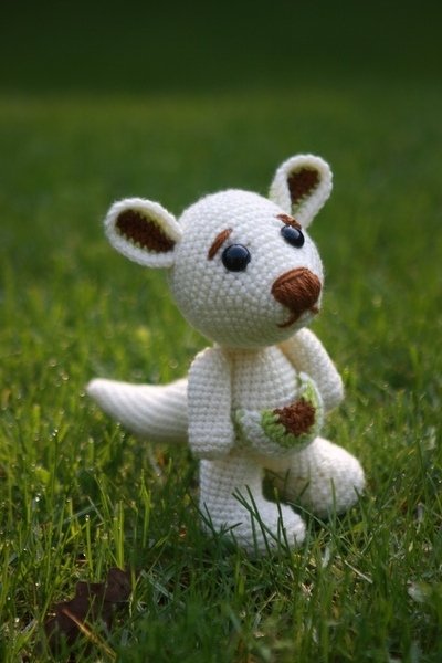 Cremeweißes gehäkeltes Känguru-Amigurumi mit braunen Ohren und Nase, sitzend im grünen Gras