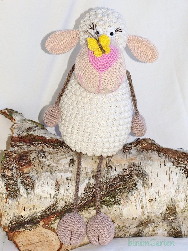 Weißes gehäkeltes Schaf-Amigurumi mit rosa Ohren und Schnauze und gelbem Schmetterling auf der Nase, sitzend auf einem Birkenholzstamm.