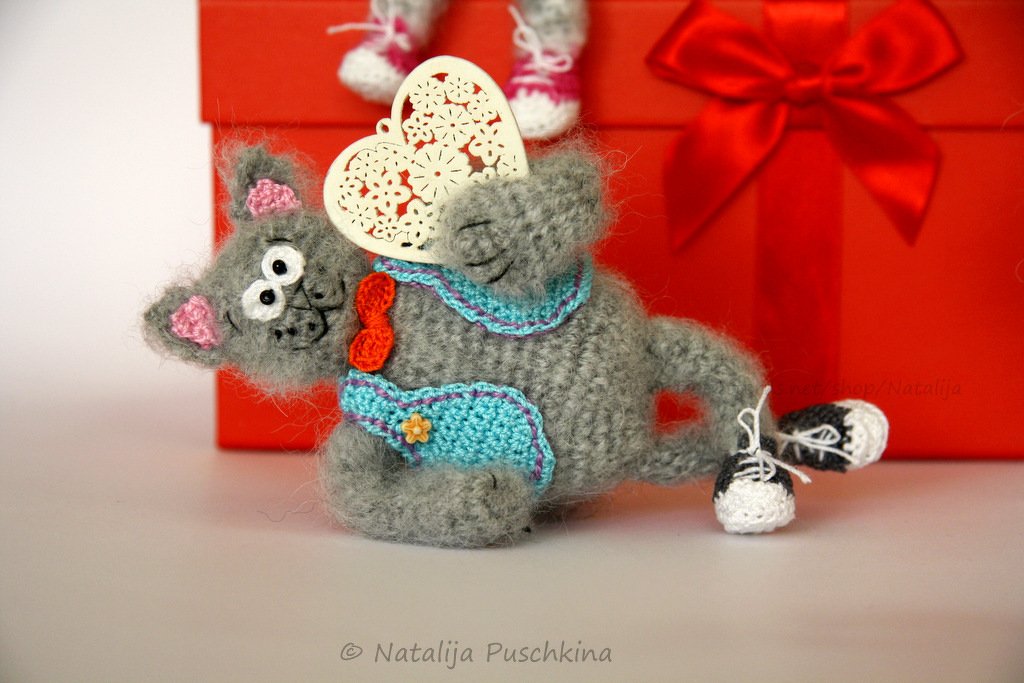 Graue gehäkelte Amigurumi-Katze mit türkisem Latz, roter Fliege, schwarz-weißen Sneakers und Herz vor roter Geschenkbox
