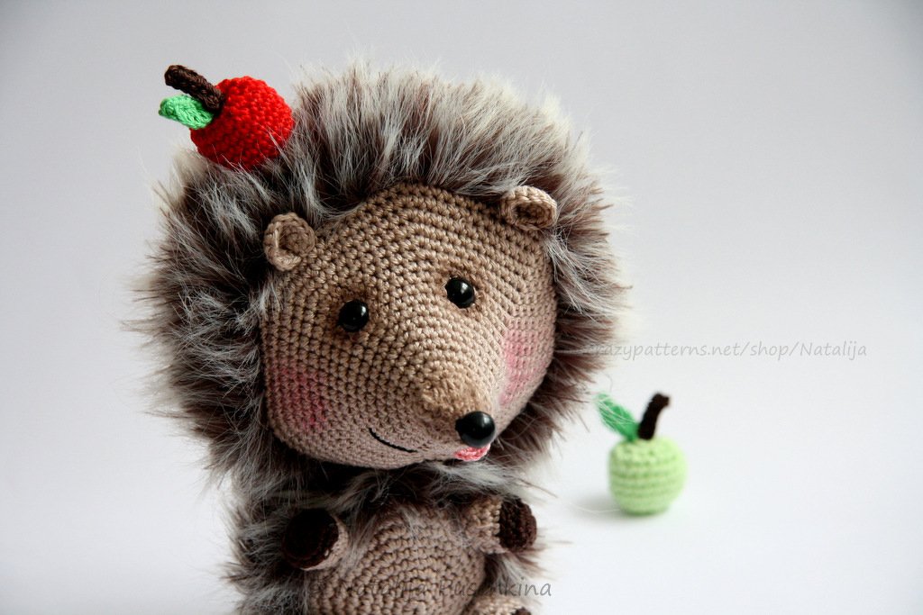 Gehäkelter Amigurumi-Igel mit flauschigen grau-braunen Stacheln und kleinem roten Apfel, grüner Apfel im Hintergrund