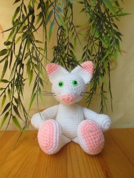 Weißes Amigurumi-Kätzchen mit rosa Ohren, rosa Nase und rosa Pfotenballen, grüne Augen, sitzt auf einem Holztisch