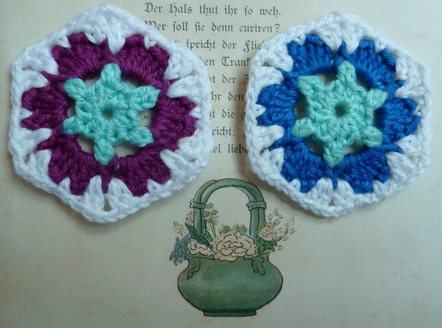 Zwei sechseckige Häkelapplikationen in Mint mit lila bzw. blauem Mittelteil und weißem Rand auf einer aufgeschlagenen Buchseite mit Illustration.