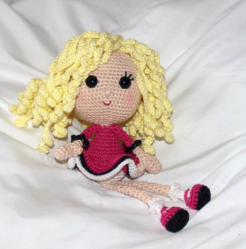 Gehäkelte Amigurumi-Puppe mit gelben Locken, pinkem Kleid und schwarzen Augen auf weißem Hintergrund