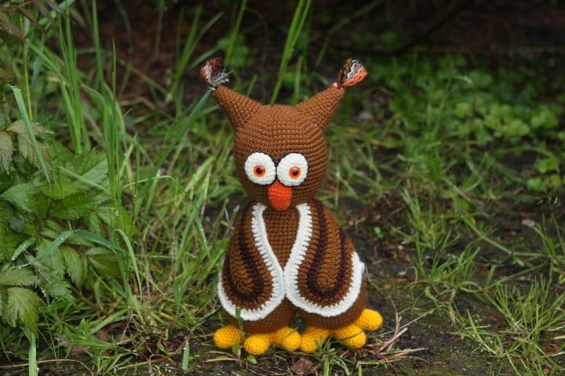 Braune handgehäkelte Amigurumi-Eule mit weißen Augenringen, orangefarbenem Schnabel und gelben Füßen auf Gras