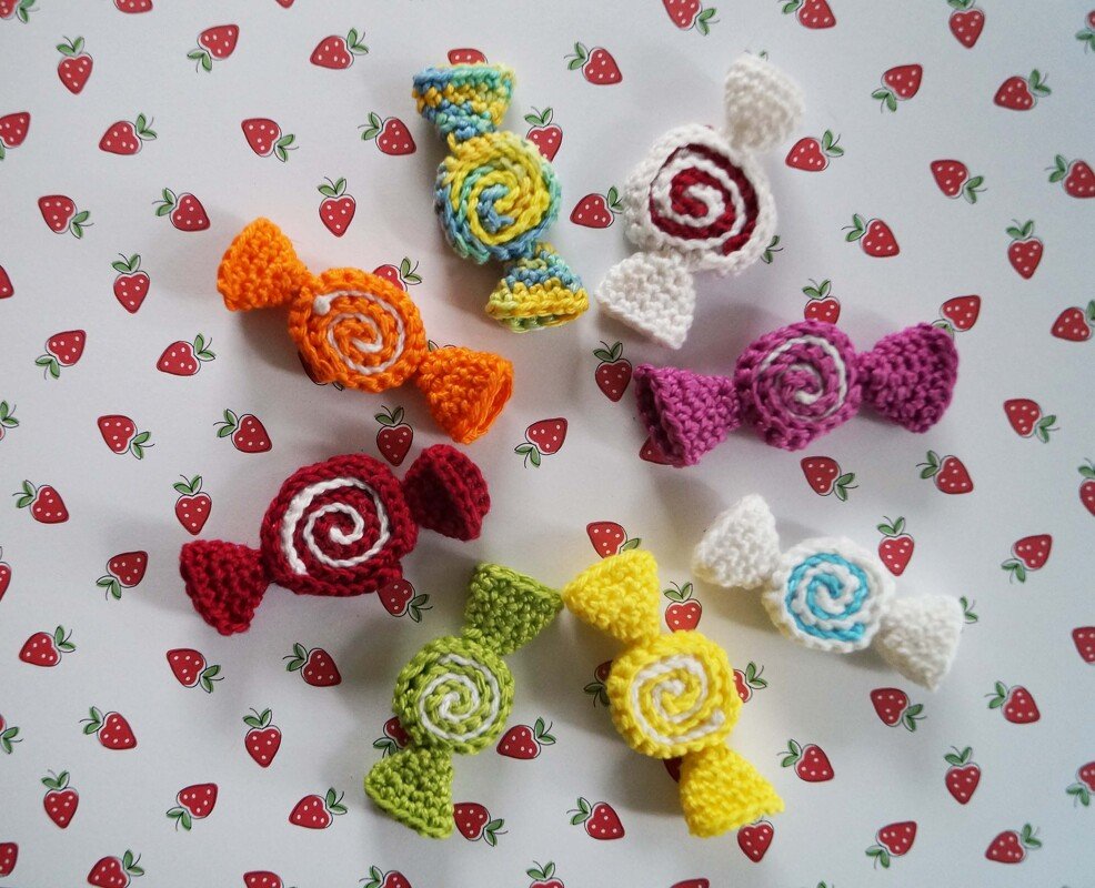 Acht gehäkelte Bonbons in verschiedenen Farben mit Spiralmuster auf einem Hintergrund mit Erdbeermuster.