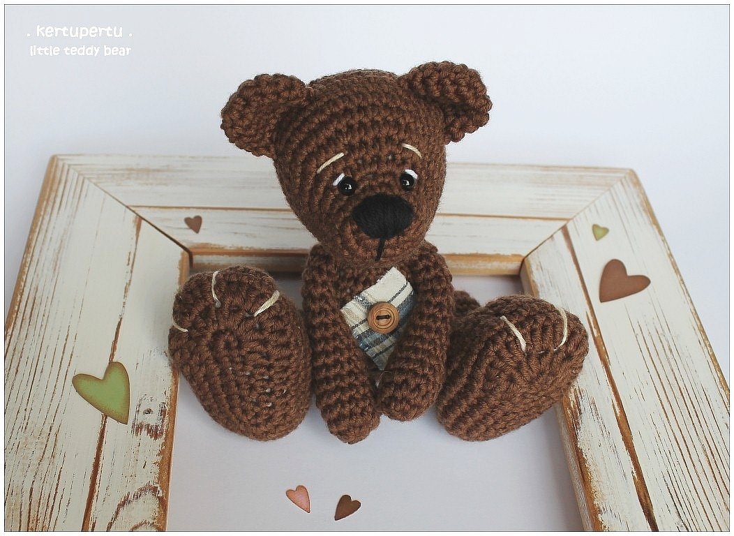 Brauner gehäkelter Amigurumi-Teddybär mit kariertem Latz und Knopf, sitzend in einem dekorativen Holzrahmen mit Herz-Aussparungen.