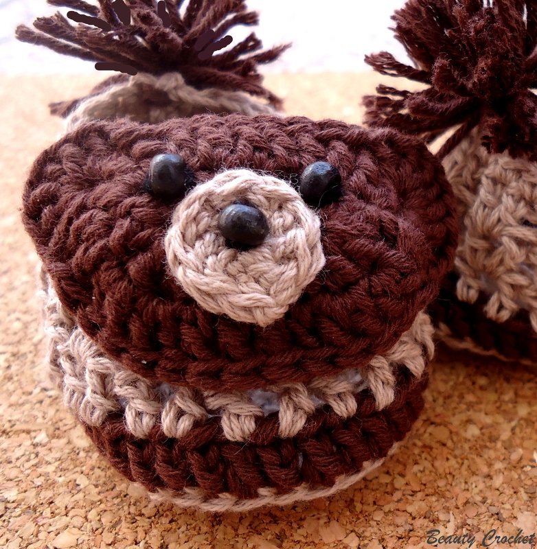 Nahaufnahme gehäkelter Babyschuhe in Braun und Beige mit Bärengesichtern und Pompons auf Korkuntergrund.