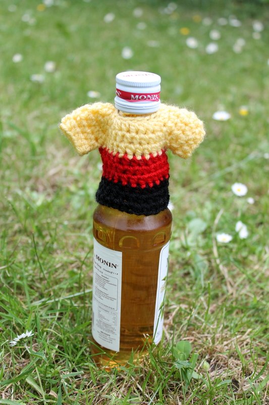 T-Shirt-förmiger Häkelüberzug in Gelb, Rot und Schwarz auf einer Flasche im Gras