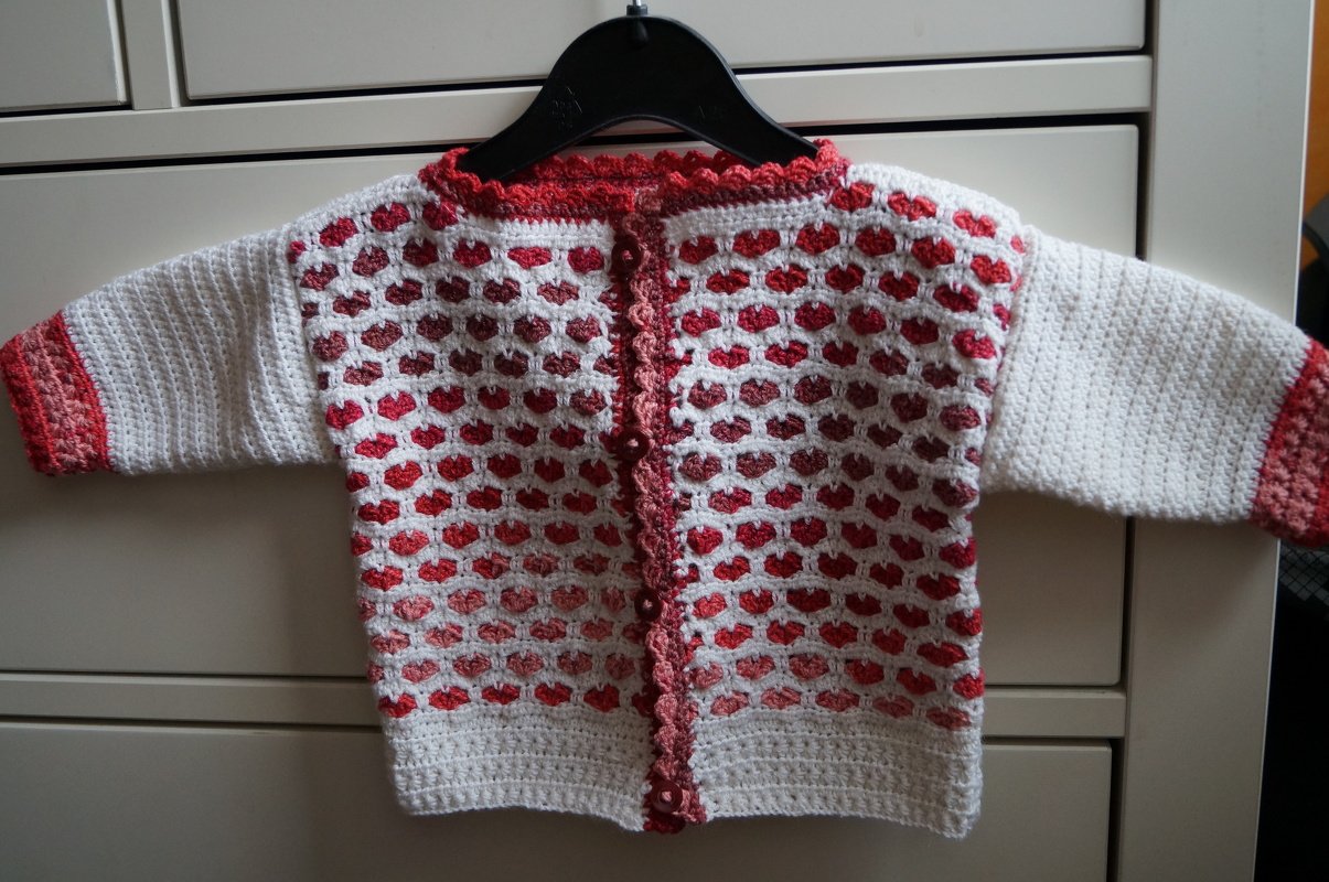 Gehäkelte Babyjacke in Weiß mit roten Herzmotiven an einem Kleiderbügel