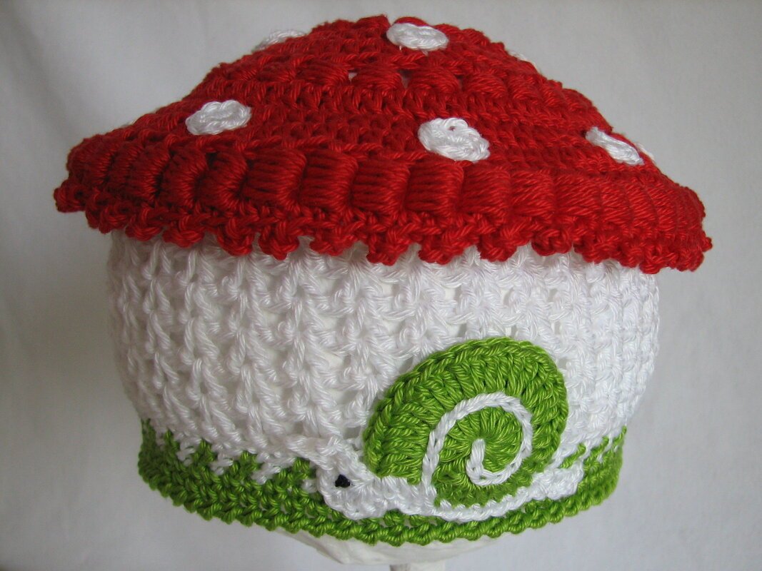 Gehäkelte Kinder‑Mütze in Pilzform mit rotem Hut, weißen Punkten und grüner Schnecke