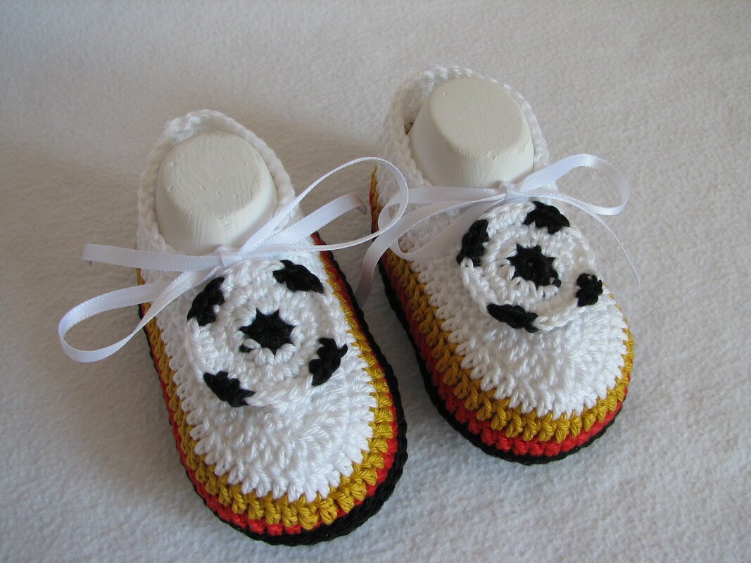 Nahaufnahme zweier weißer, gehäkelter Babyschuhe mit schwarz‑rot‑goldenem Rand und kleinen Fußball‑Applikationen, verziert mit weißen Schleifenbändern.