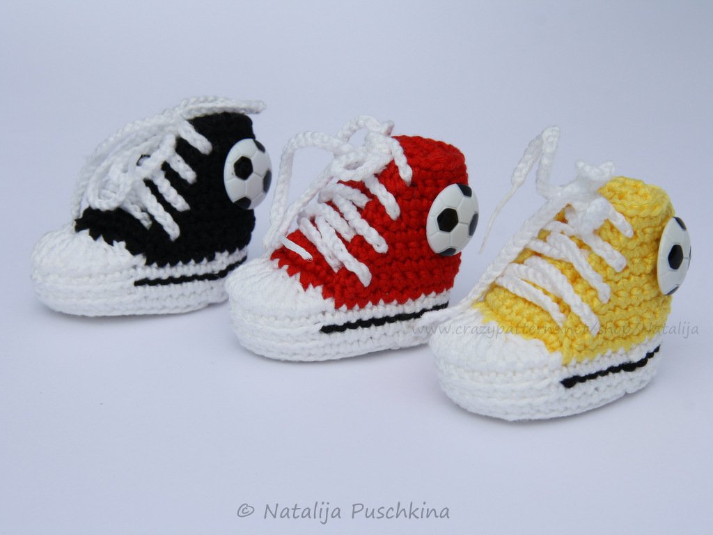 Drei gehäkelte Baby-Sneaker in Schwarz, Rot und Gelb mit weißen Schnürsenkeln und Fußball-Knöpfen