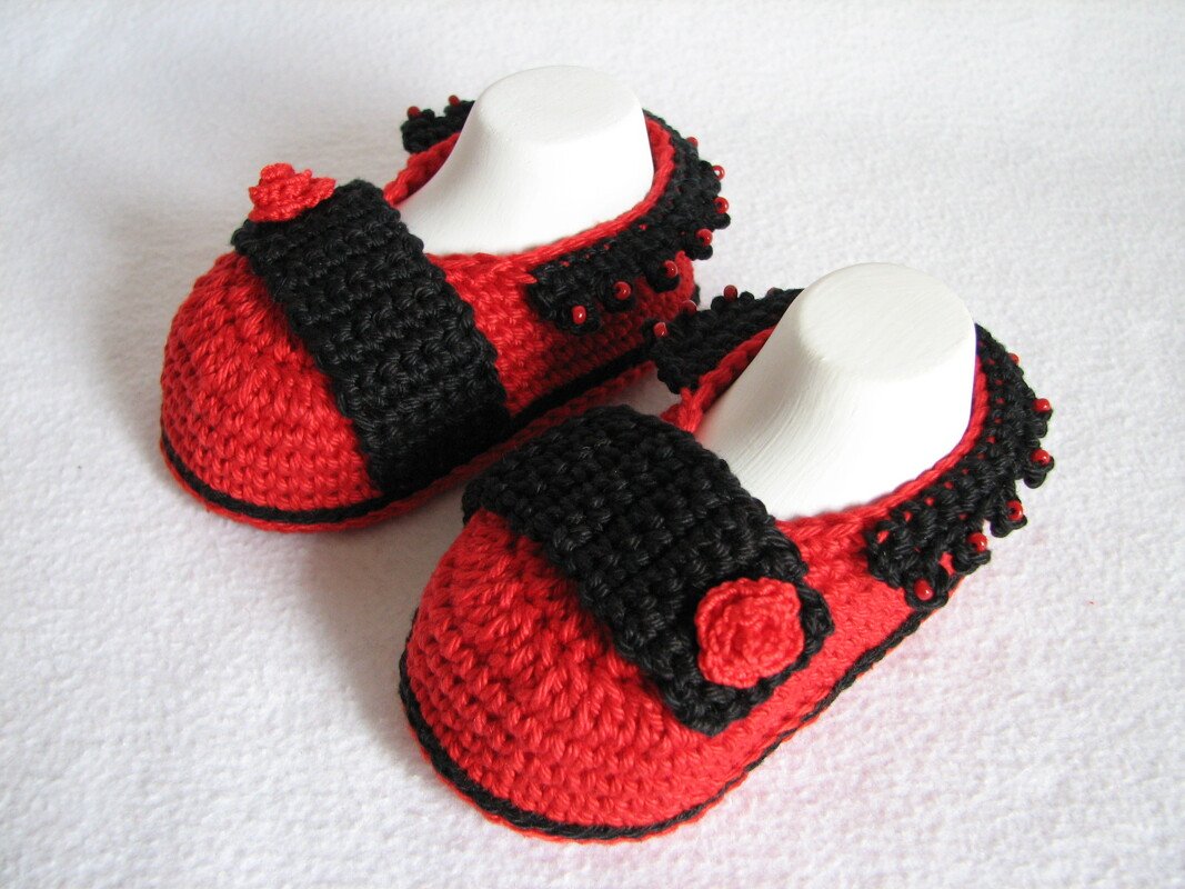 Rote und schwarze gehäkelte Babyschuhe mit schwarzem Riemen, roter Blüte und Perlen auf weißem Hintergrund.