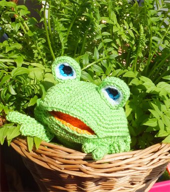 Gehäkelter Amigurumi-Frosch mit blauen Augen und oranger Mundöffnung, sitzend in einem Weidenkorb.