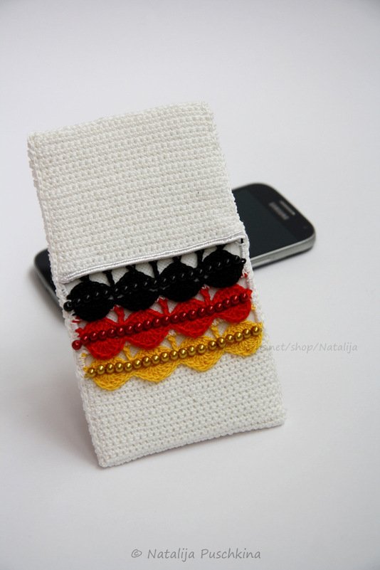 Weiße gehäkelte Handytasche mit schwarz-rot-gelben Muschelreihen und Perlenstickerei.