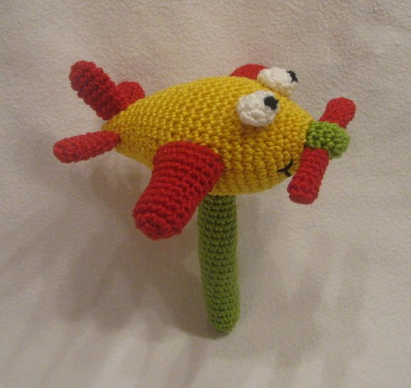 Gehäkelte Babyrassel in Form eines gelben Flugzeugs mit roten Flügeln, grünem Griff und Propeller.