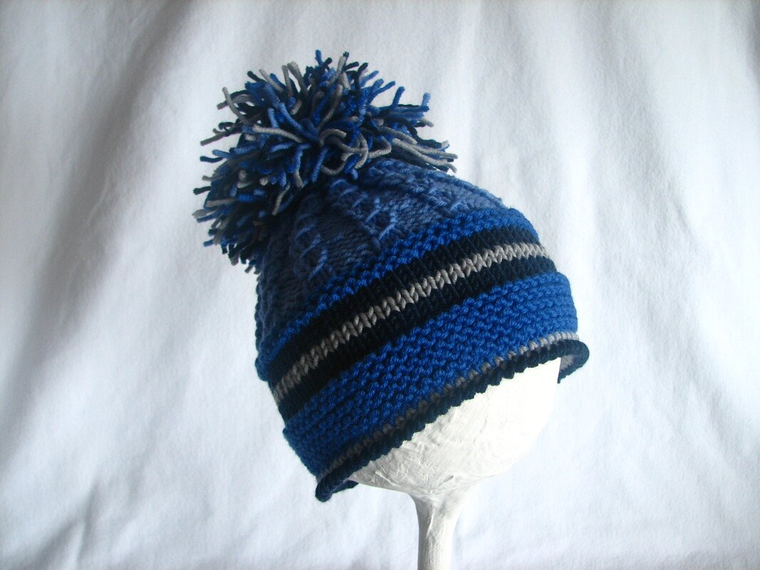 Gestrickte Kindermütze mit Bommel in Blau- und Grautönen auf weißem Ständer
