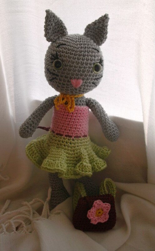 Grau gehäkelte Amigurumi-Katze mit rosa Oberteil, grünem Rüschenrock und kleiner Tasche