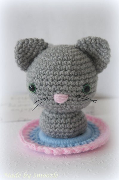 Gehäkelter grauer Amigurumi-Mäusekopf mit rosa Nase und grünen Augen auf runder Unterlage