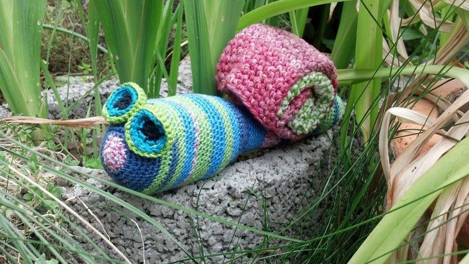 Gehäkelte Amigurumi‑Schnecke mit blau‑grünem Streifenkörper und rosa‑grünem Schneckenhaus auf Stein im Gras
