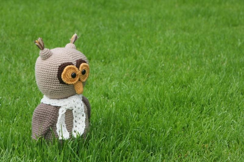 Gehäkelte Amigurumi-Eule in Braun mit weißem Schal auf grünem Rasen.