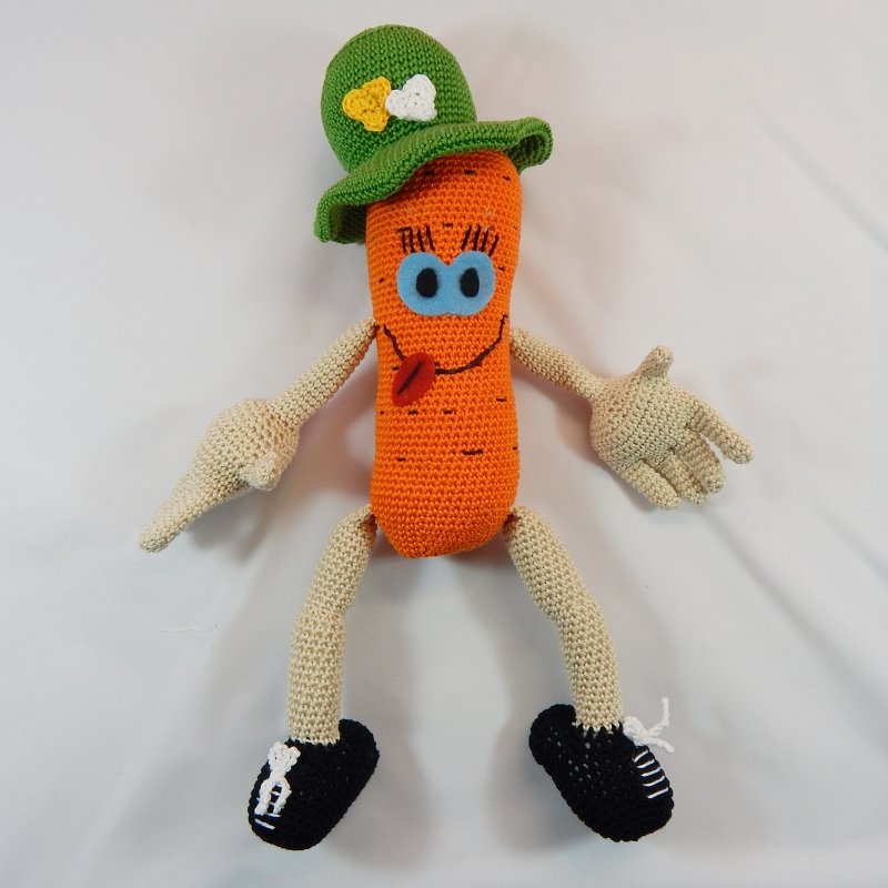 Gehäkelte orange Karottenfigur mit blauen Augen, roter Zunge, grünem Hut und schwarzen Schuhen auf weißem Hintergrund.