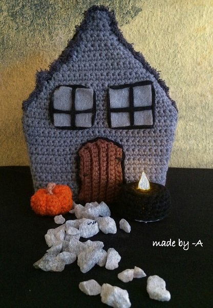 Gehäkeltes Spukhaus in Grau mit Fenster- und Türdetails, Amigurumi‑Kürbis, LED‑Teelicht und weißen Dekosteinen