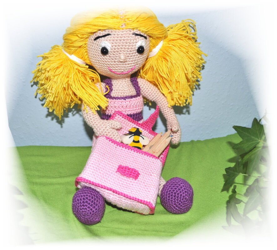 Handgehäkelte Puppe mit gelbem Haar, rosa Tasche und Holzstiften auf grünem Stoff.