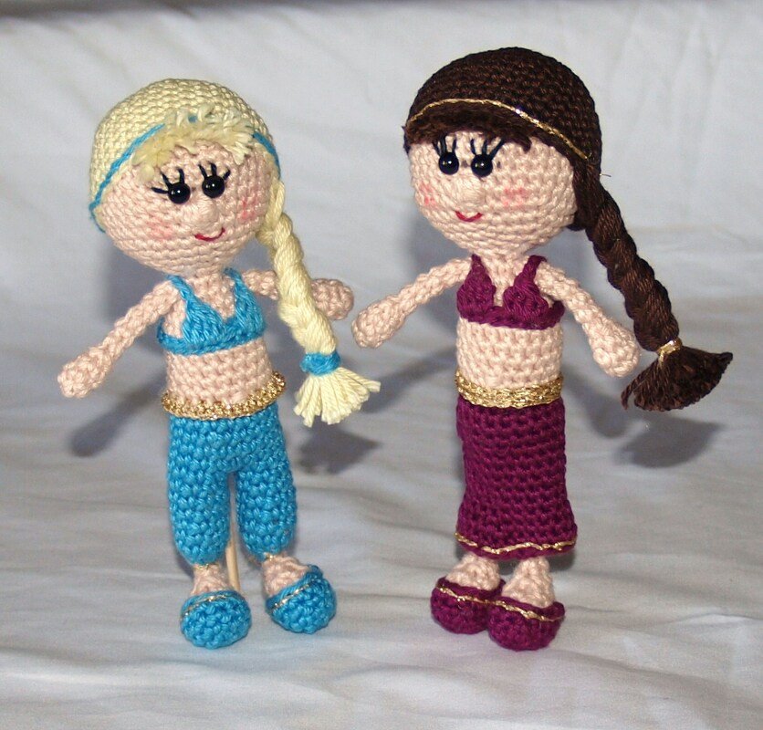 Zwei gehäkelte Amigurumi-Puppen mit geflochtenen Zöpfen, blauem Hosen-Outfit, violettem Kleid und goldenen Bändern