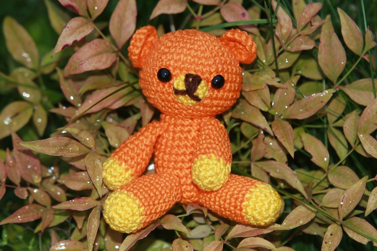 Nahaufnahme eines sitzenden gehäkelten Teddybären in Orange mit gelben Pfoten