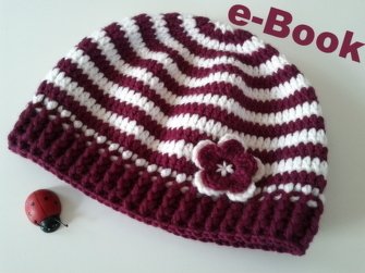 Gehäkelte Babymütze mit dunkelrot-weißen Ringelstreifen, geripptem Bund und einer kleinen Blume, daneben eine Marienkäfer-Figur.
