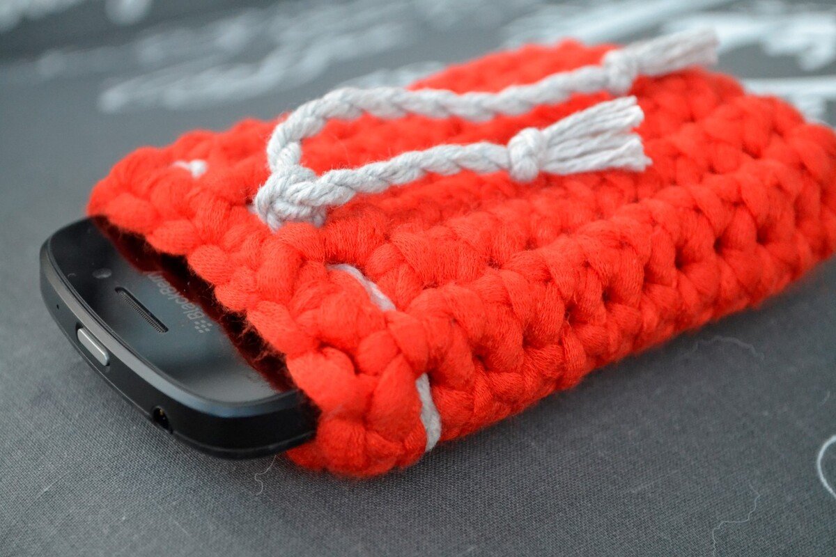 Rote, grob gehäkelte Handyhülle mit grauem Zugband und teilweise eingestecktem Smartphone