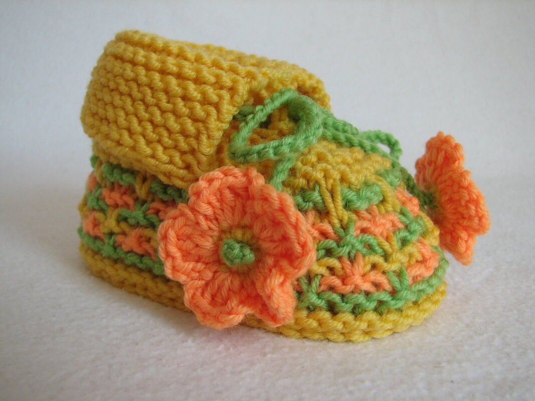 Gestrickter Babyschuh in Gelb, Orange und Grün mit orangefarbener Blume und grünem Band auf hellem Hintergrund.