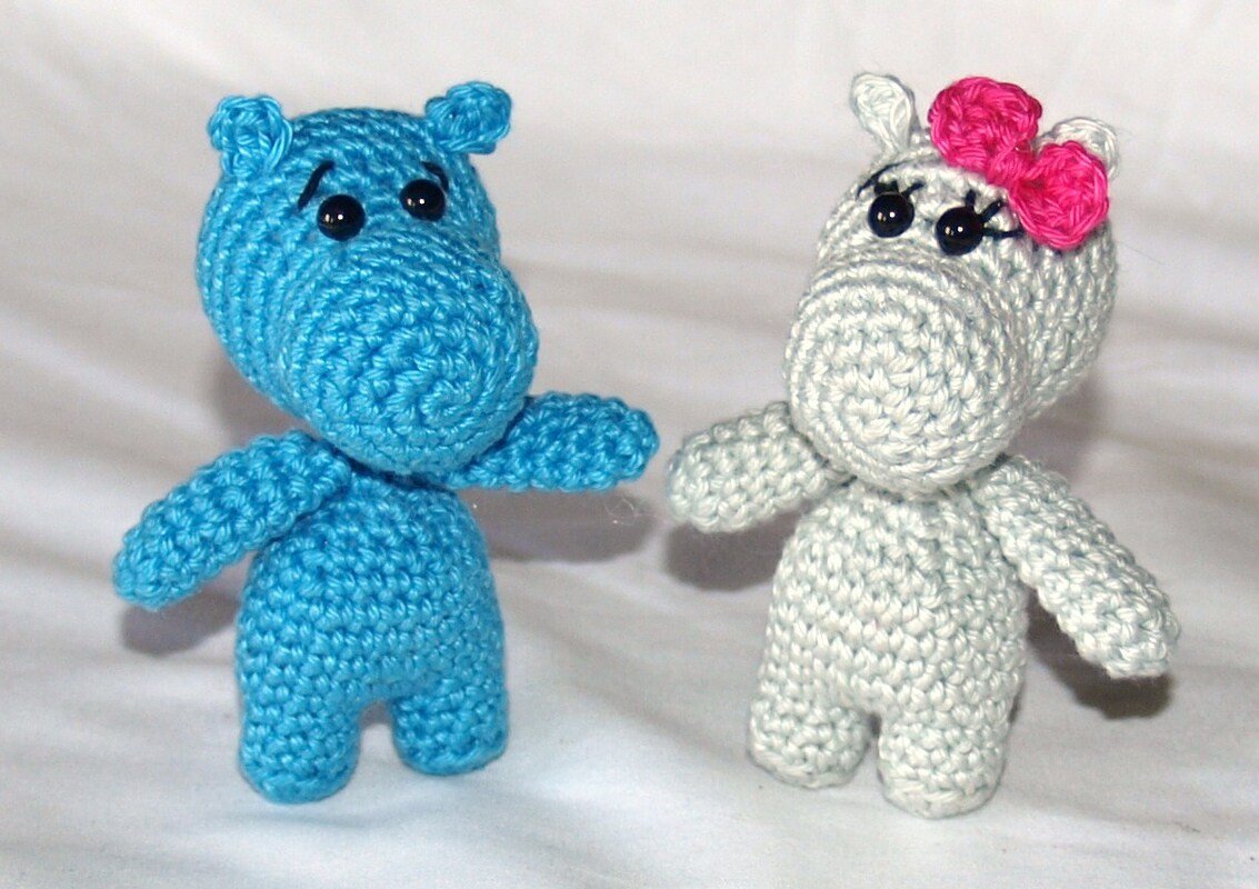 Zwei gehäkelte Amigurumi-Nilpferde, eines blau und eines hellgrau mit rosa Schleife