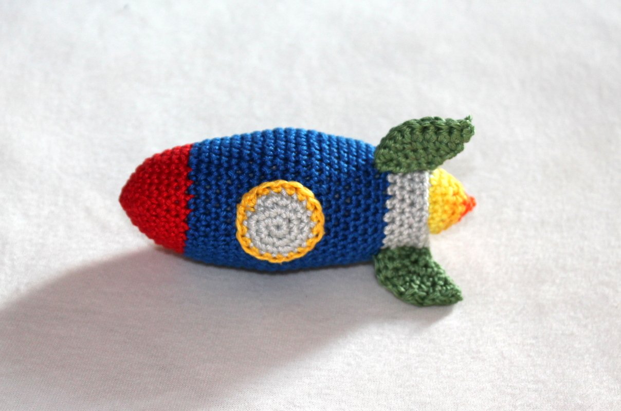 Gehäkelte, bunte Amigurumi-Rakete liegend auf hellem Stoff