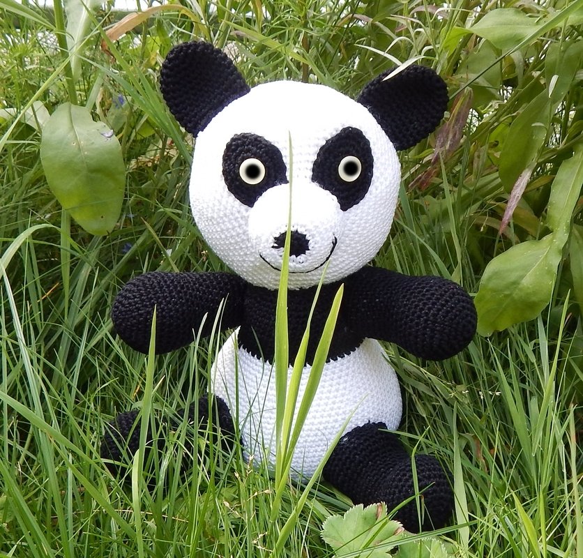Schwarz-weißer gehäkelter Amigurumi-Panda sitzt im hohen grünen Gras.